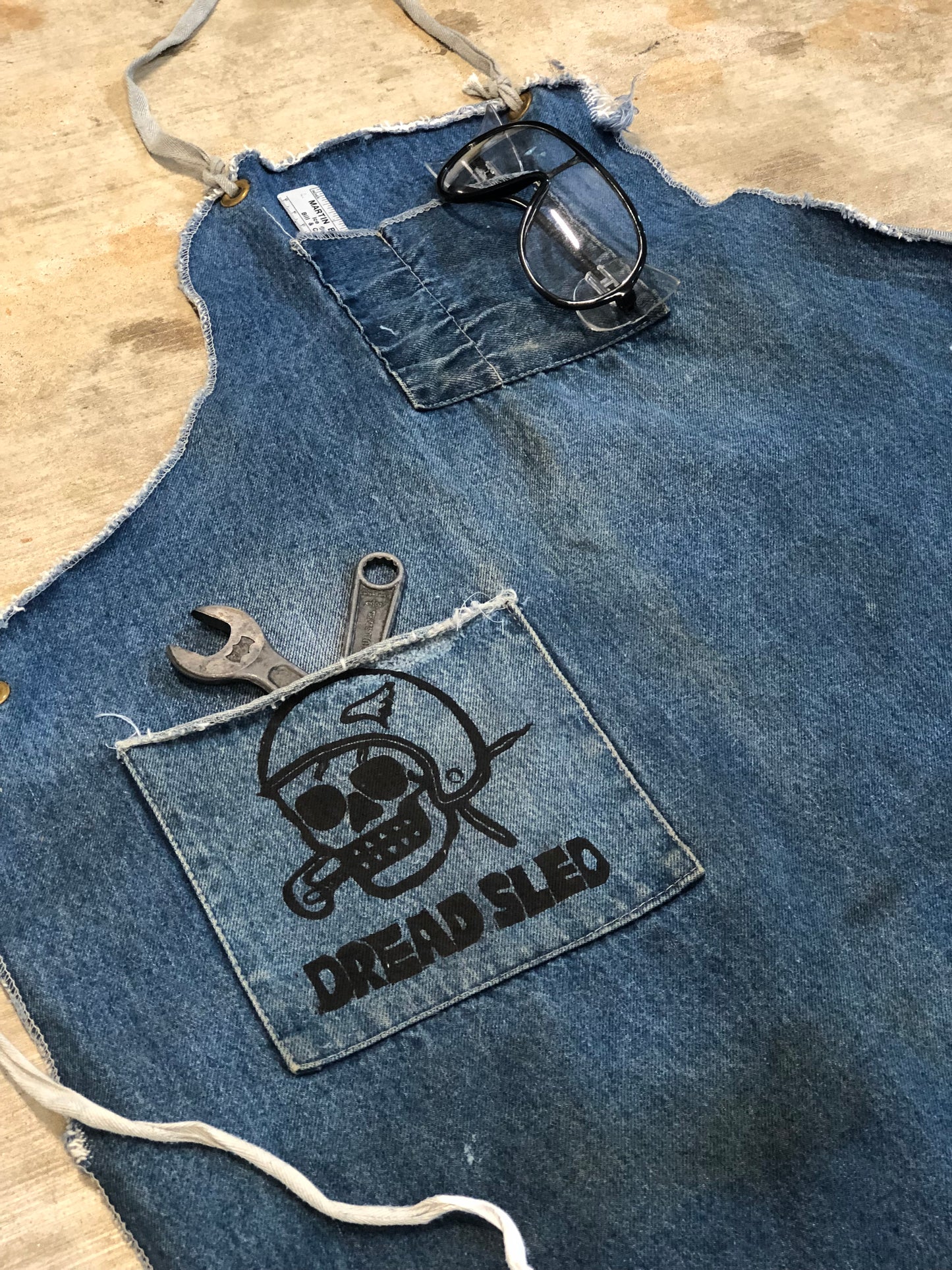 Blue denim apron with a logo patch and tools on a concrete surface